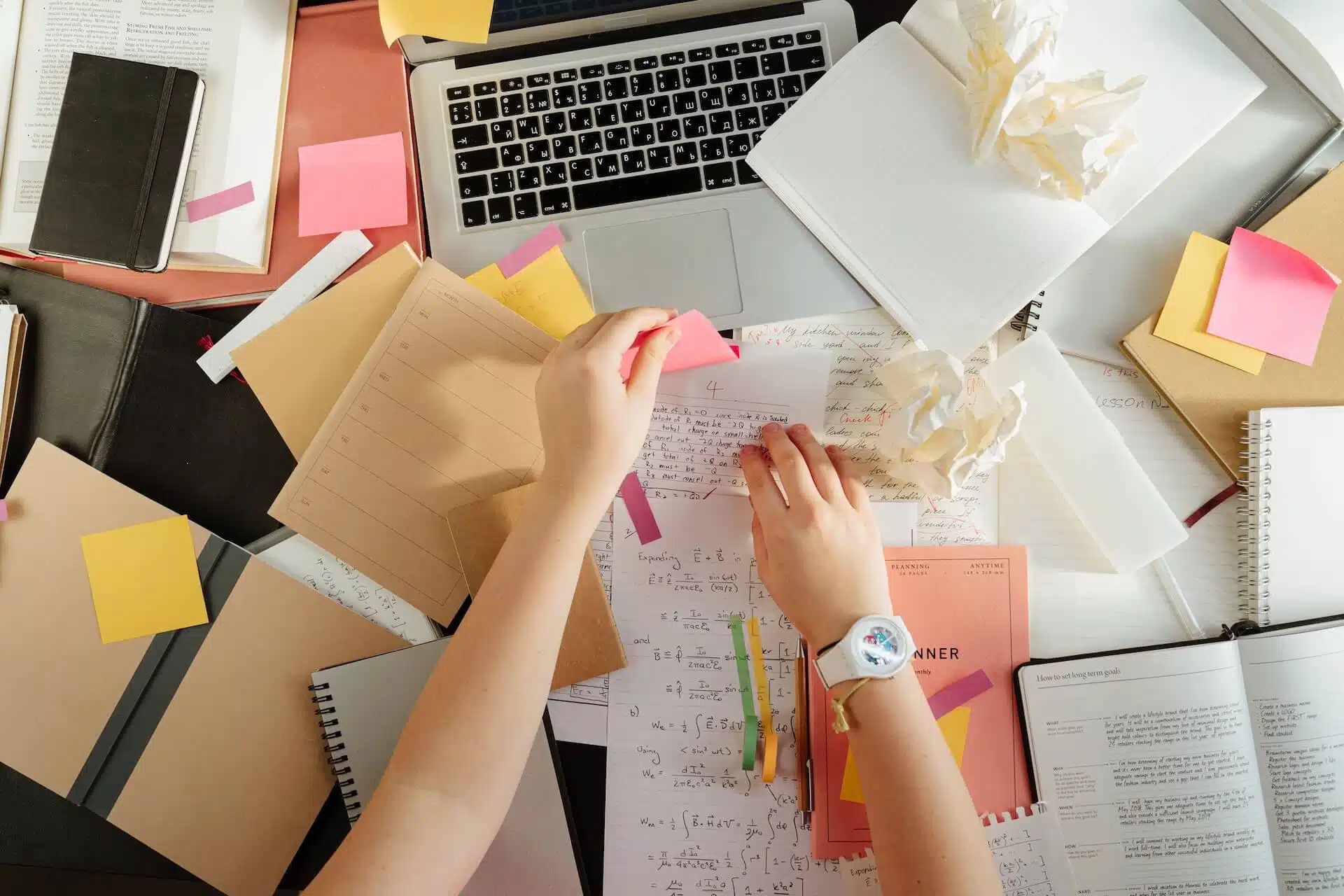 Mani su una scrivania disordinata piena di libri, appunti e post-it, tema della procrastinazione